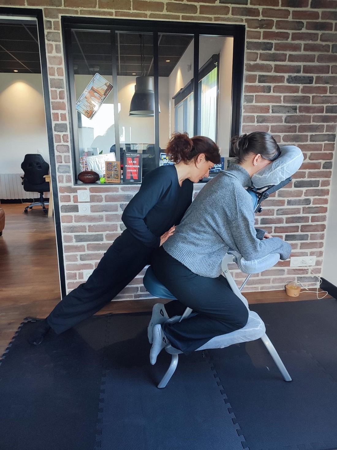 Solène Canivet en intervention de shiatsu sur chaise dans un espace professionnel — mur de briques apparentes et verrière industrielle